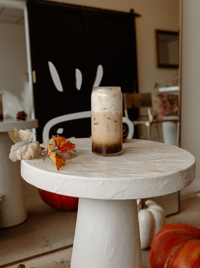 An iced latte on a white pedestal in front of the smiling Latte Haus mascot wall, with pumpkins